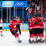 Team Canada women's ice hockey players in red and white jerseys huddle together in celebration after scoring a goal against Team Switzerland. A scoreboard in the background shows Canada leading 1-0 during the 2026 Winter Olympics match, with fans cheering in the stands holding Canadian flags.
