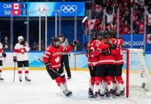 Team Canada women's ice hockey players in red and white jerseys huddle together in celebration after scoring a goal against Team Switzerland. A scoreboard in the background shows Canada leading 1-0 during the 2026 Winter Olympics match, with fans cheering in the stands holding Canadian flags.