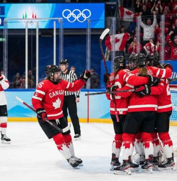 Team Canada women's ice hockey players in red and white jerseys huddle together in celebration after scoring a goal against Team Switzerland. A scoreboard in the background shows Canada leading 1-0 during the 2026 Winter Olympics match, with fans cheering in the stands holding Canadian flags.
