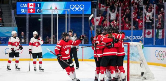 Team Canada women's ice hockey players in red and white jerseys huddle together in celebration after scoring a goal against Team Switzerland. A scoreboard in the background shows Canada leading 1-0 during the 2026 Winter Olympics match, with fans cheering in the stands holding Canadian flags.