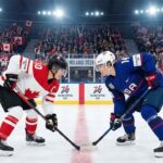 Team Canada captain Marie-Philip Poulin (left, #29) and Team USA forward Hilary Knight (right, #21) are crouched and ready for a face-off at center ice during a women's ice hockey game at the Milano Cortina 2026 Winter Olympics. The arena is filled with spectators waving Canadian and American flags, and banners reading "MILANO 2026" are visible on the boards.