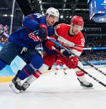 Auston Matthews (USA) and Nikolaj Ehlers (Denmark) battle intensely for the puck along the boards with Olympic branding visible during the Milano Cortina 2026 Men's Ice Hockey tournament.