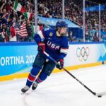 A Team USA women's ice hockey player wearing a blue number 28 jersey skates with the puck along the boards marked "Milano Cortina 2026" and Olympic rings during a Winter Olympic game, with fans in the background.