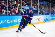 A Team USA women's ice hockey player wearing a blue number 28 jersey skates with the puck along the boards marked "Milano Cortina 2026" and Olympic rings during a Winter Olympic game, with fans in the background.