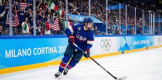 A Team USA women's ice hockey player wearing a blue number 28 jersey skates with the puck along the boards marked "Milano Cortina 2026" and Olympic rings during a Winter Olympic game, with fans in the background.