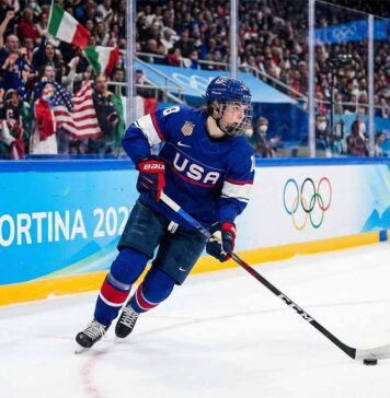 A Team USA women's ice hockey player wearing a blue number 28 jersey skates with the puck along the boards marked "Milano Cortina 2026" and Olympic rings during a Winter Olympic game, with fans in the background.
