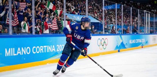 A Team USA women's ice hockey player wearing a blue number 28 jersey skates with the puck along the boards marked "Milano Cortina 2026" and Olympic rings during a Winter Olympic game, with fans in the background.