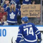 Toronto Maple Leafs winger Bobby McMann (#74) kneels on the ice facing the crowd, where a smiling female fan behind the glass wears a Leafs toque and jersey, holding up a handmade cardboard sign that reads, "Good luck with your new team Bobby!"