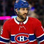 Montreal Canadiens center Vincent Trocheck, wearing his red No. 16 home jersey and a blue helmet with a visor, looks towards the right side of the rink. The background shows blurred spectators and distinct magenta and blue arena lights.