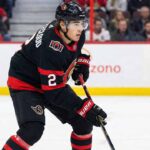 Zach Whitecloud of the Ottawa Senators, wearing a black uniform with the number 2, stands on the ice holding his hockey stick and looking forward during a game.