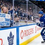 Toronto Maple Leafs fan behind glass at a hockey game holding a hand-drawn sign that says 'PLEASE DON'T LEAVE TORONTO AUSTON!' directed at Auston Matthews (#34) who is skating past on the ice.