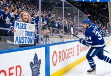 Toronto Maple Leafs fan behind glass at a hockey game holding a hand-drawn sign that says 'PLEASE DON'T LEAVE TORONTO AUSTON!' directed at Auston Matthews (#34) who is skating past on the ice.