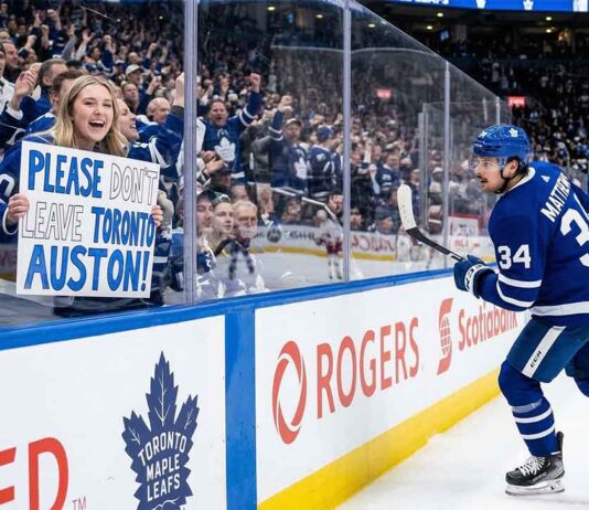 Toronto Maple Leafs fan behind glass at a hockey game holding a hand-drawn sign that says 'PLEASE DON'T LEAVE TORONTO AUSTON!' directed at Auston Matthews (#34) who is skating past on the ice.