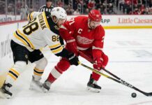 Boston Bruins David Pastrnak and Detroit Red Wings Dylan Larkin battle intensely for a puck near the boards at Little Caesars Arena during an Atlantic Division matchup.