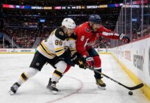 An intense puck battle along the boards between Boston Bruins forward David Pastrnak (88) in a white away jersey and Washington Capitals captain Alex Ovechkin (8) in a red home jersey during an NHL game at Capital One Arena.
