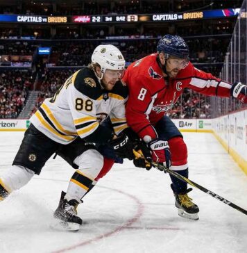 An intense puck battle along the boards between Boston Bruins forward David Pastrnak (88) in a white away jersey and Washington Capitals captain Alex Ovechkin (8) in a red home jersey during an NHL game at Capital One Arena.