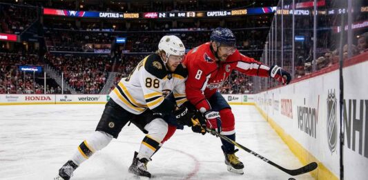 An intense puck battle along the boards between Boston Bruins forward David Pastrnak (88) in a white away jersey and Washington Capitals captain Alex Ovechkin (8) in a red home jersey during an NHL game at Capital One Arena.