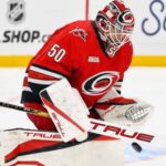 Why the Carolina Hurricanes Passed on a Jordan Binnington Trade A conceptual image showing St. Louis Blues goaltender Jordan Binnington wearing a full red Carolina Hurricanes uniform and goalie gear, in a blocking pose on the ice.