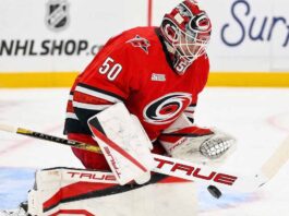 Why the Carolina Hurricanes Passed on a Jordan Binnington Trade A conceptual image showing St. Louis Blues goaltender Jordan Binnington wearing a full red Carolina Hurricanes uniform and goalie gear, in a blocking pose on the ice.