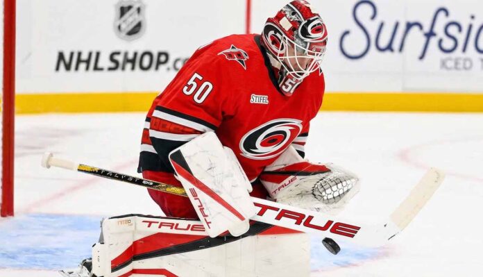 A conceptual image showing St. Louis Blues goaltender Jordan Binnington wearing a full red Carolina Hurricanes uniform and goalie gear, in a blocking pose on the ice.
