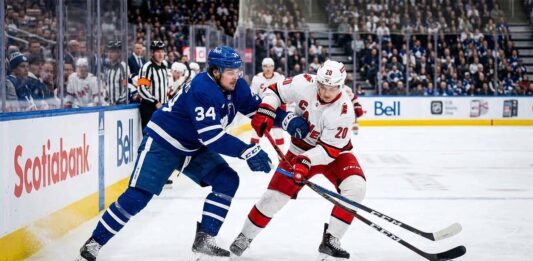 Leafs Face Ultimate Test Against Hurricanes After The Devastating Matthews Injury Action shot of Auston Matthews #34 (Toronto Maple Leafs) and Sebastian Aho #20 (Carolina Hurricanes) engaged in a close battle for puck possession on the ice during an NHL game.