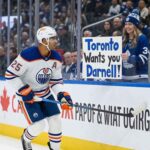 Edmonton Oilers defenseman Darnell Nurse #25 skating past a fan holding a trade sign, as rumors heat up about his $9.25M contract.