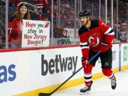 Dougie Hamilton of the New Jersey Devils skates near the boards while a fan holds a sign reading "Hope you Stay in New Jersey Dougie!" amidst offseason trade rumors.