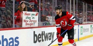 Dougie Hamilton of the New Jersey Devils skates near the boards while a fan holds a sign reading "Hope you Stay in New Jersey Dougie!" amidst offseason trade rumors.