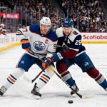 Edmonton Oilers captain Connor McDavid (#97) in an away white jersey battles Colorado Avalanche alternate captain Nathan MacKinnon (#29) for the puck during a high-stakes NHL game.