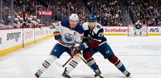 Edmonton Oilers captain Connor McDavid (#97) in an away white jersey battles Colorado Avalanche alternate captain Nathan MacKinnon (#29) for the puck during a high-stakes NHL game.