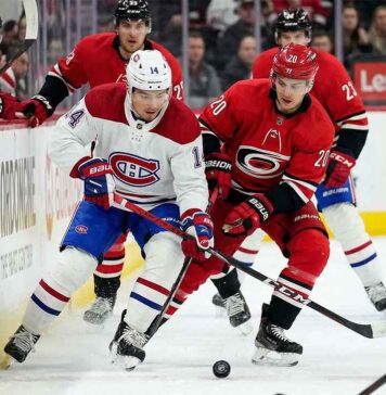 Montreal Canadiens captain Nick Suzuki (number 14) in white battles Carolina Hurricanes center Sebastian Aho (number 20) in red for puck control during an NHL game. The Lenovo Center logo is visible on the boards in the background.