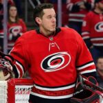 Carolina Hurricanes Trade Rumors: Jordan Binnington & St. Louis Blues A conceptual photograph of St. Louis Blues goaltender Jordan Binnington, wearing a detailed Carolina Hurricanes red goalie uniform with the spiral logo, leaning against a red goal post.