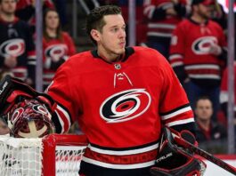 Carolina Hurricanes Trade Rumors: Jordan Binnington & St. Louis Blues A conceptual photograph of St. Louis Blues goaltender Jordan Binnington, wearing a detailed Carolina Hurricanes red goalie uniform with the spiral logo, leaning against a red goal post.