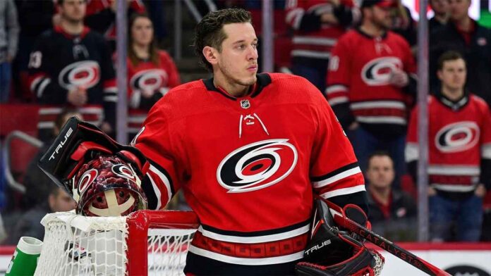 A conceptual photograph of St. Louis Blues goaltender Jordan Binnington, wearing a detailed Carolina Hurricanes red goalie uniform with the spiral logo, leaning against a red goal post.