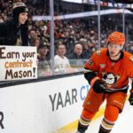 Mason McTavish Trade Rumors: Will the Anaheim Ducks Move Him? Anaheim Ducks center Mason McTavish skates along the boards past a fan holding a sign reading "EARN YOUR CONTRACT MASON!" highlighting the pressure on the young forward amidst trade rumors.