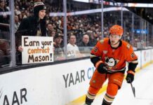 Anaheim Ducks center Mason McTavish skates along the boards past a fan holding a sign reading "EARN YOUR CONTRACT MASON!" highlighting the pressure on the young forward amidst trade rumors.