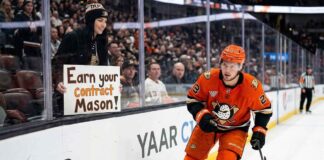 Anaheim Ducks center Mason McTavish skates along the boards past a fan holding a sign reading "EARN YOUR CONTRACT MASON!" highlighting the pressure on the young forward amidst trade rumors.