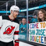 New Jersey Devils defenseman Dougie Hamilton looks at a hand-painted sign held by a smiling San Jose Sharks fan through the arena glass. The sign reads 'COME TO SAN JOSE DOUGIE!', referencing player movement and trade rumors between the two teams.