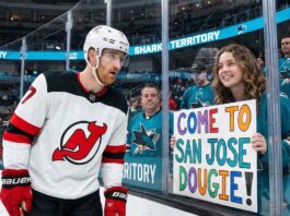 New Jersey Devils defenseman Dougie Hamilton looks at a hand-painted sign held by a smiling San Jose Sharks fan through the arena glass. The sign reads 'COME TO SAN JOSE DOUGIE!', referencing player movement and trade rumors between the two teams.