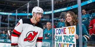 New Jersey Devils defenseman Dougie Hamilton looks at a hand-painted sign held by a smiling San Jose Sharks fan through the arena glass. The sign reads 'COME TO SAN JOSE DOUGIE!', referencing player movement and trade rumors between the two teams.