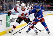 Close-up action photograph capturing Ottawa Senators' captain Brady Tkachuk (#7) and New York Rangers' center Vincent Trocheck (#16) intensely battling for the puck during an NHL game, with a blurred background of spectators and arena boards.