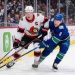 Ottawa Senators captain Brady Tkachuk (number 7 in white jersey) battles a Vancouver Canucks player (number 6 in blue jersey) for the puck during an NHL game along the boards at Rogers Arena.