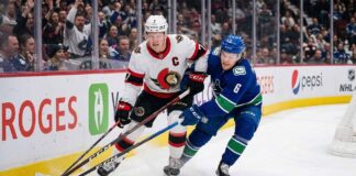 Ottawa Senators captain Brady Tkachuk (number 7 in white jersey) battles a Vancouver Canucks player (number 6 in blue jersey) for the puck during an NHL game along the boards at Rogers Arena.