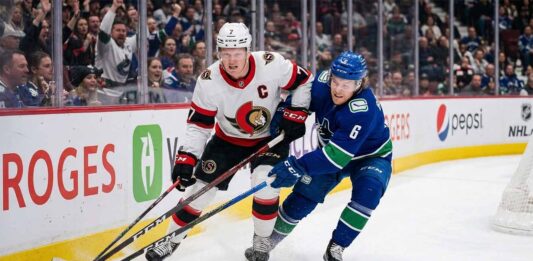 Ottawa Senators captain Brady Tkachuk (number 7 in white jersey) battles a Vancouver Canucks player (number 6 in blue jersey) for the puck during an NHL game along the boards at Rogers Arena.