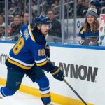 St. Louis Blues center Robert Thomas #18 in white jersey skating along the boards as a female fan holds a sign that reads "Please stay in St. Louis Robert!" amidst NHL trade rumors connecting him to the Buffalo Sabres and Utah Mammoth.