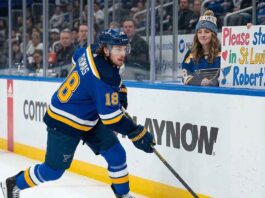 St. Louis Blues center Robert Thomas #18 in white jersey skating along the boards as a female fan holds a sign that reads "Please stay in St. Louis Robert!" amidst NHL trade rumors connecting him to the Buffalo Sabres and Utah Mammoth.