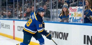 St. Louis Blues center Robert Thomas #18 in white jersey skating along the boards as a female fan holds a sign that reads "Please stay in St. Louis Robert!" amidst NHL trade rumors connecting him to the Buffalo Sabres and Utah Mammoth.