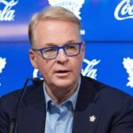 Toronto Maple Leafs Fire GM Brad Treliving – Search for Data-Centric GM MLSE President and CEO Keith Pelley speaking at a press conference about the Toronto Maple Leafs seeking a new data-centric GM. He is wearing distinctive blue-rimmed glasses and sitting in front of a blue backdrop with Maple Leafs logos and Coca-Cola branding.