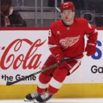 Will the Detroit Red Wings Trade Alex DeBrincat This Offseason? Detroit Red Wings forward Alex DeBrincat skating on the ice, wearing the red winged-wheel jersey, with Coca-Cola and Samsung advertisements on the rink boards.