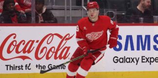 Detroit Red Wings forward Alex DeBrincat skating on the ice, wearing the red winged-wheel jersey, with Coca-Cola and Samsung advertisements on the rink boards.